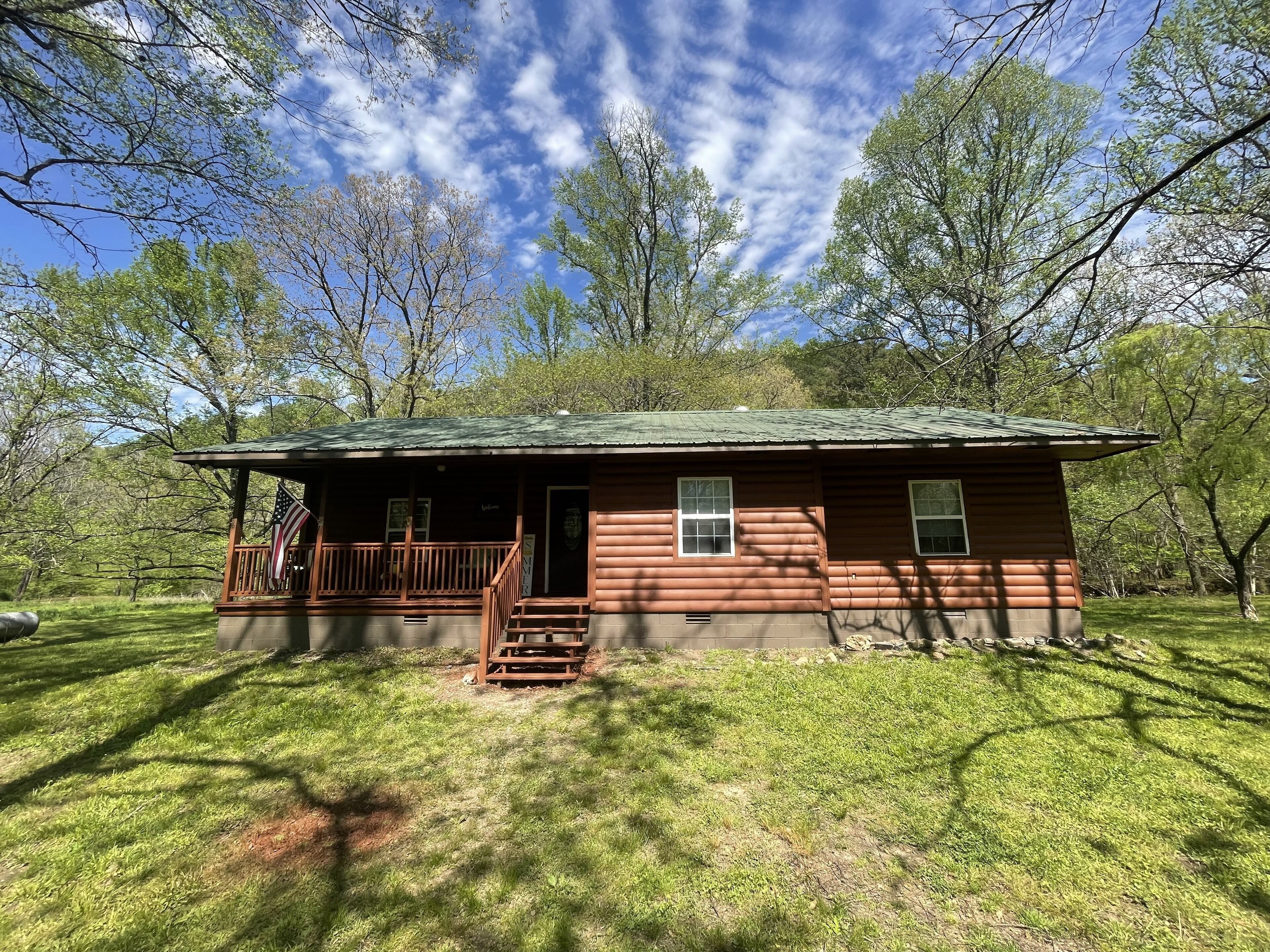 A small wooden cabin with a porch, surrounded by trees and grass.