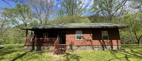 A small wooden cabin with a porch, surrounded by trees and grass.