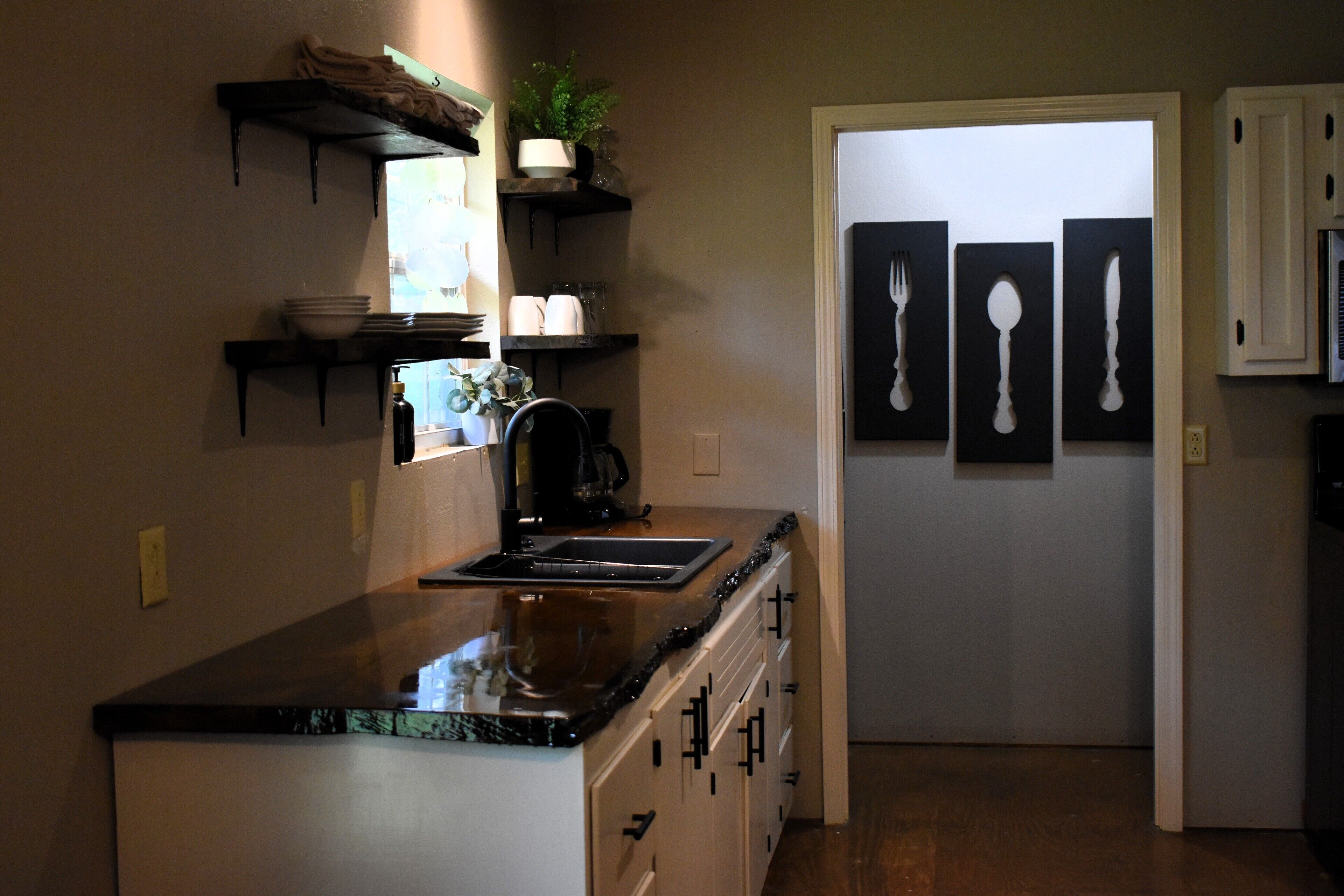 Una cocina con encimera negra, armarios blancos y una cafetera. Hay estantes sobre la encimera con vajilla y una planta.