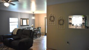 A living room with a dark leather sofa, a dining area with a table and chairs, and a kitchen visible through an open doorway. - Peaceful Cabin Steps Away From The Creek, Convenient Access To ATV Trails! (Mena)