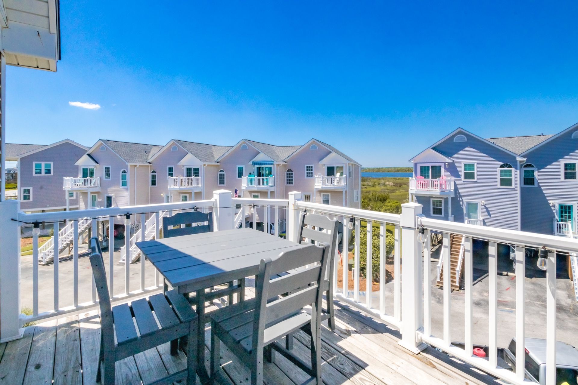 A wooden deck with a table and chairs, overlooking a row of houses with balconies, under a clear blue sky.