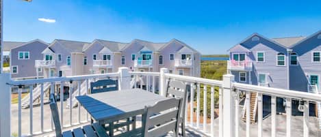 A wooden deck with a table and chairs, overlooking a row of houses with balconies, under a clear blue sky.