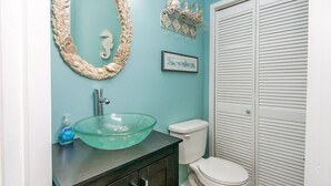 A bathroom with a glass sink, a toilet, a framed picture, and a decorative mirror.