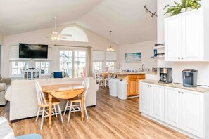 A spacious living area with a wooden dining table, white sofas, and a kitchen with white cabinets and a coffee maker.