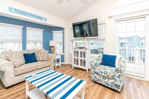 A living room with a sofa, armchair, and a coffee table. There is a TV mounted on the wall and a window with blinds. - Followin' The Son Too - 3 BR in Calinda Cay; Comm. Pool & Ocean Views (North Topsail Beach)