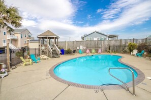 A swimming pool with a wooden deck, a gazebo, and a fenced backyard with colorful chairs.