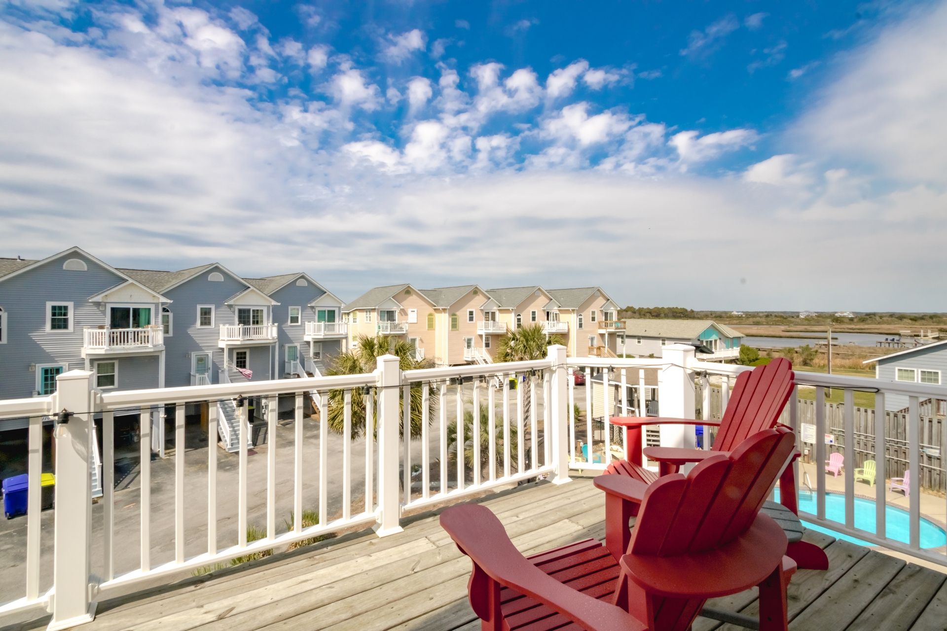 Una terraza de madera con sillas Adirondack rojas que dan a una hilera de casas de playa con balcones.