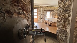 A stone-walled bathroom with a shower and a view into a bedroom with a bed and a wooden bench. - The Stone House (Kakodiki)