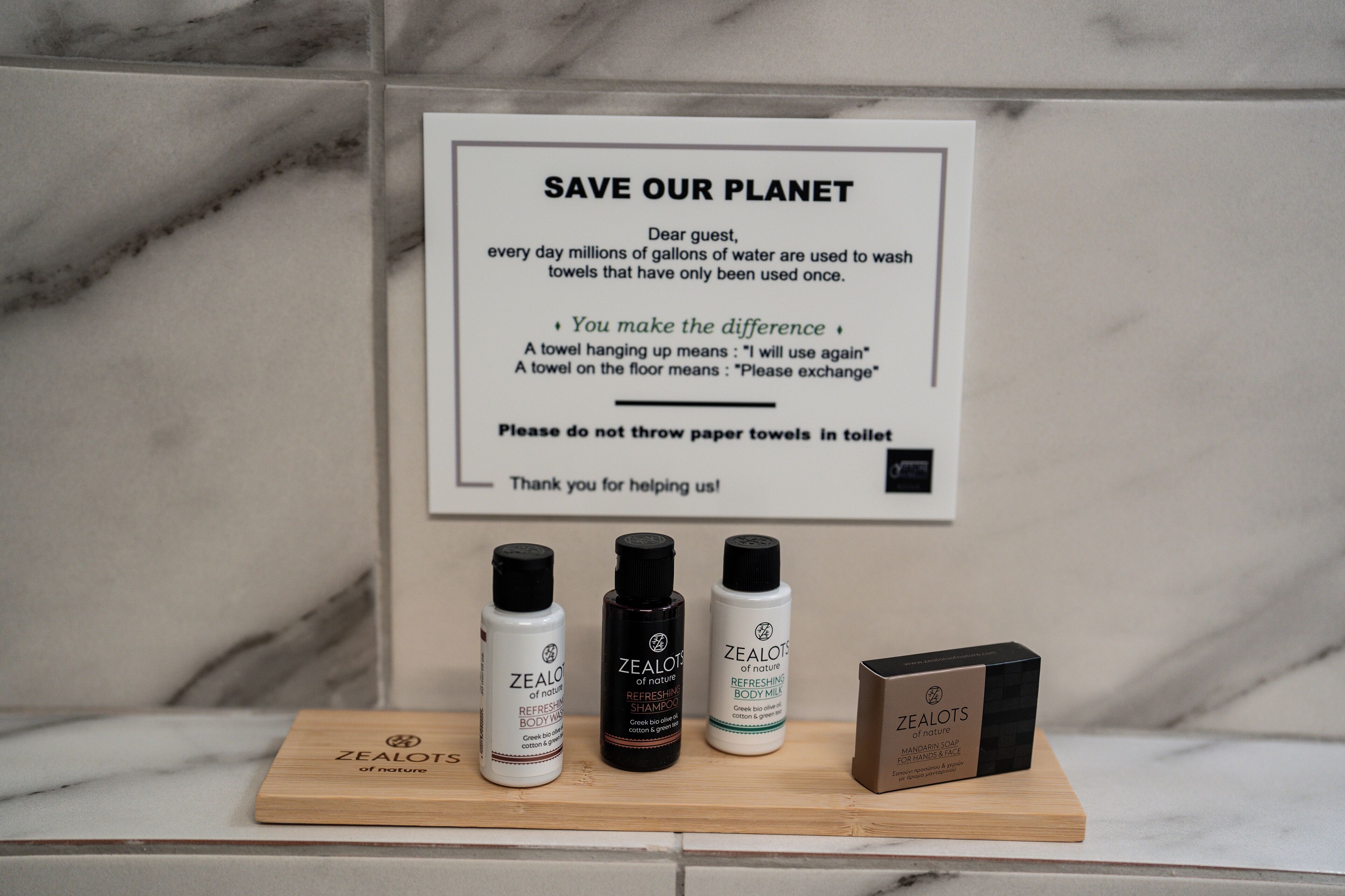 Une salle de bain avec une pancarte encourageant à réutiliser les serviettes, trois bouteilles de produits de la marque Zealots et une boîte en bois.