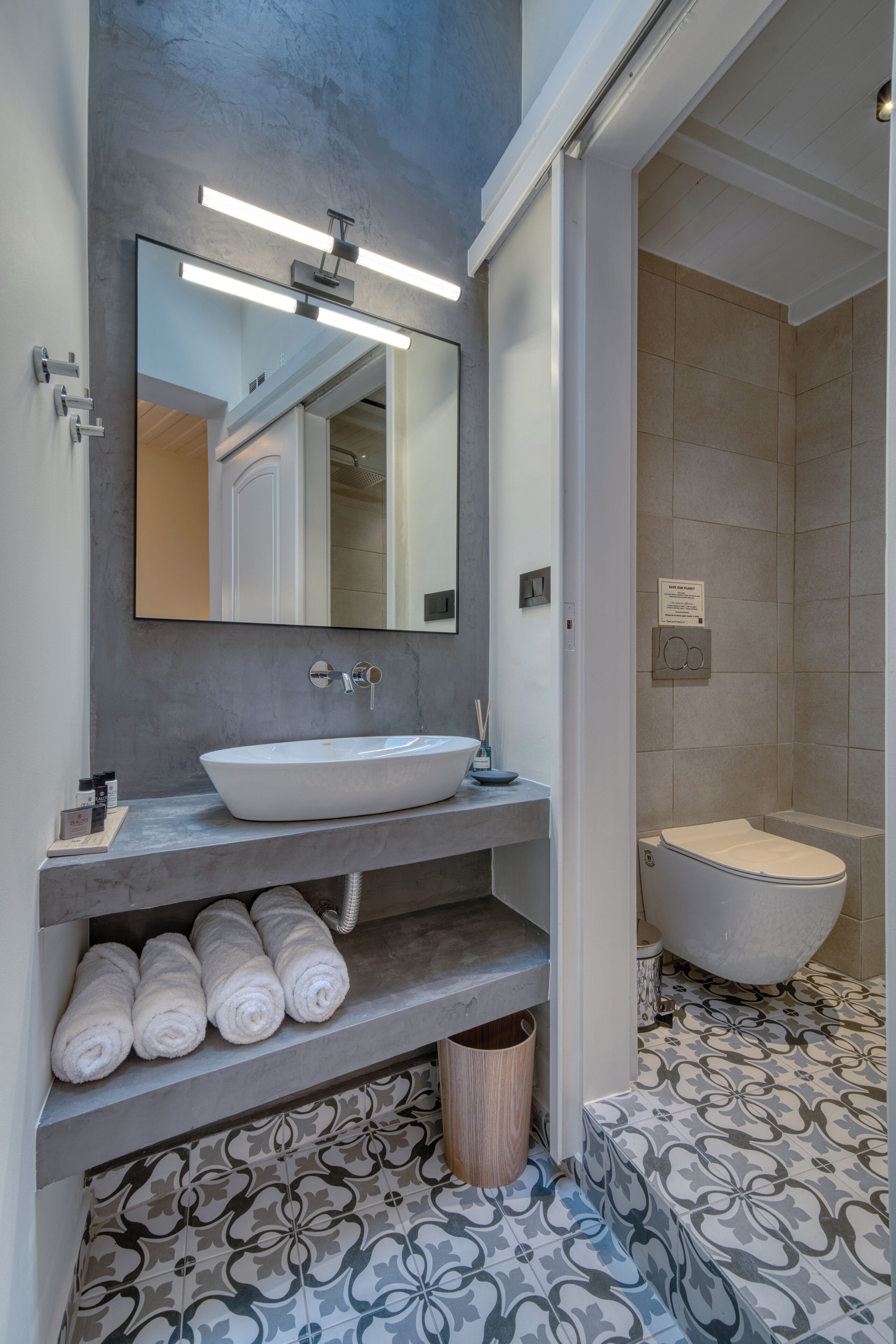 A modern bathroom with a large mirror, a white sink, and a toilet. There are towels on a shelf and a patterned floor.
