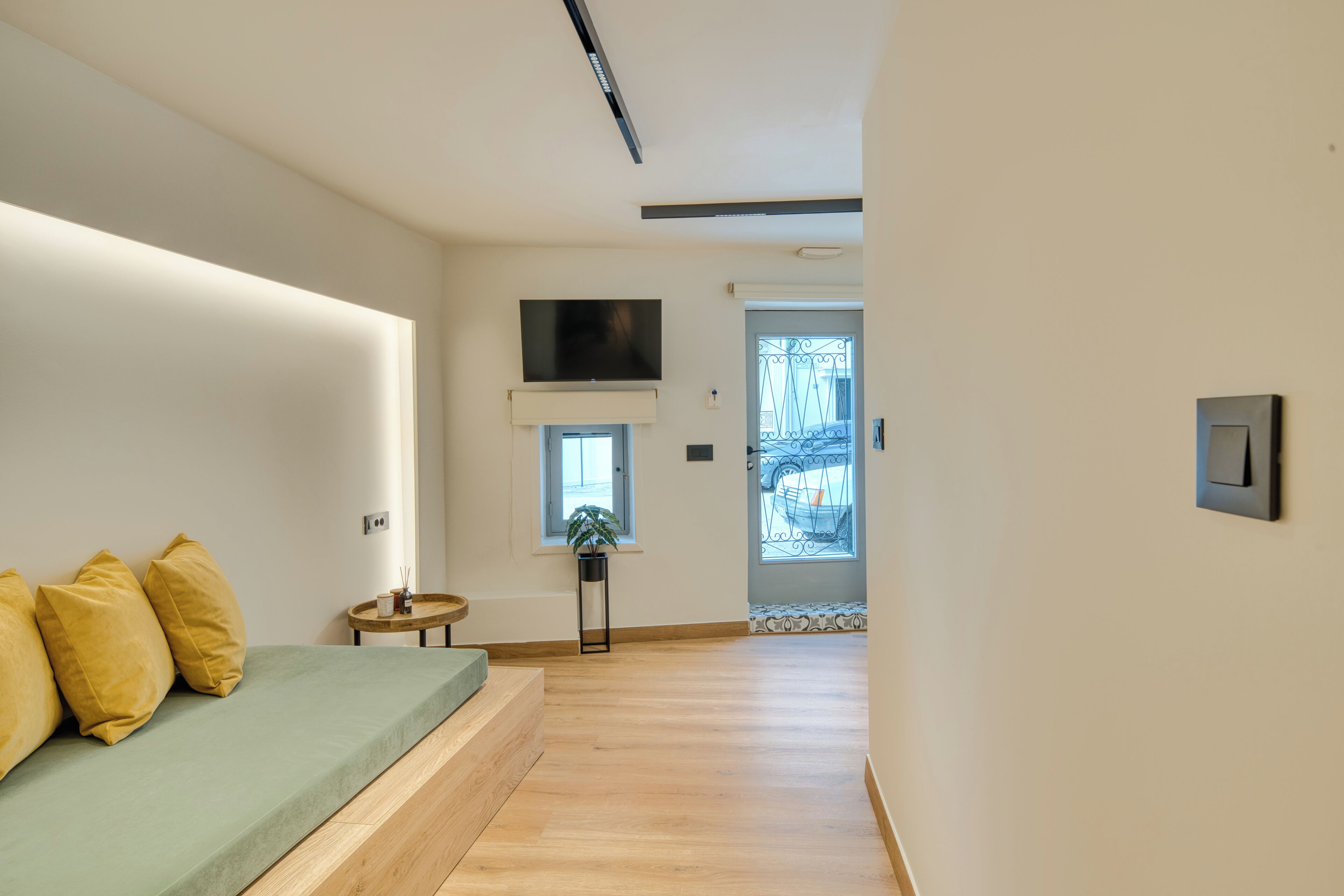 Una habitación de hotel moderna con piso de madera, un sofá verde con cojines amarillos, una pequeña mesa redonda, una planta en maceta y un televisor de pantalla plana montado en la pared.