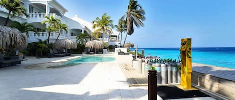 Área de piscina do resort com vista clara do oceano, posto de salva-vidas e um edifício branco com varandas.
