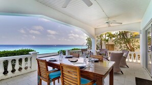 Un coin repas avec vue sur l’océan, des chaises en bois, une table dressée pour deux personnes et un ventilateur de plafond.