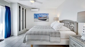 Une chambre à coucher avec un lit, un ventilateur de plafond, un tableau représentant un bateau et une fenêtre avec des rideaux.
