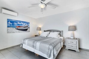 A bedroom with a bed, two bedside tables, a ceiling fan, an air conditioning unit, and a painting of a boat on the wall. - Belmar Oceanfront Apartment 1, sea views (Kralendijk)