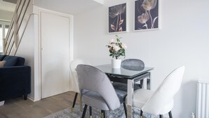 A modern dining area with a glass table, two grey chairs, a white vase with flowers, and two framed floral artworks on the wall. - Luxurious 2-bed House in Dartford (Dartford)