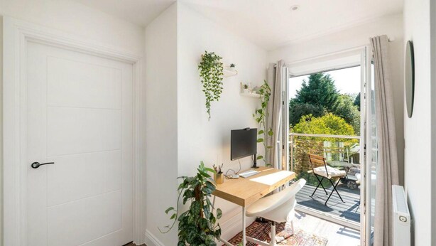 A room with a white door, a desk with a computer, a chair, and a plant. There is a balcony with a view of trees and a chair outside. - 2 Bed 2 Bath Apt Close to Reigate Station - 2230 (Reigate)