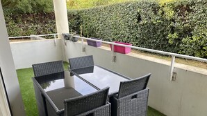 Un balcon doté d’une table à plateau en verre et de quatre chaises en osier, entouré de gazon synthétique et d’une haie.