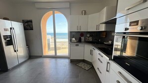 Een moderne keuken met uitzicht op zee, met een koelkast, oven en witte kastjes.