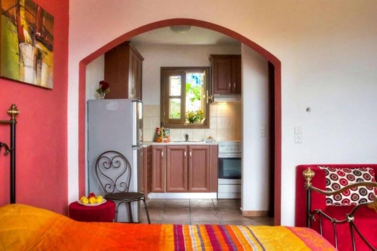 A bedroom with a bed, a chair, and a small table with fruit. There is a kitchen area with cabinets, a refrigerator, and a window.