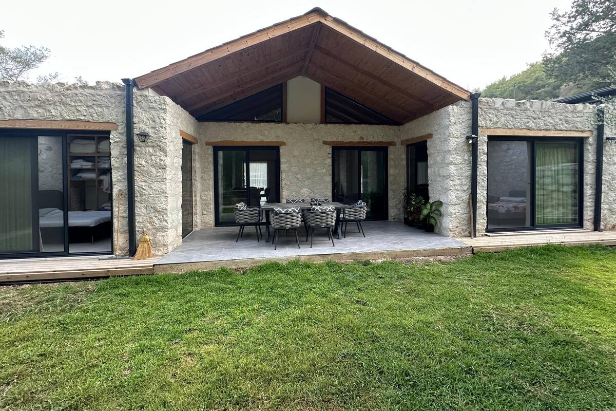 Ein modernes Haus mit Natursteinfassade, einer überdachten Terrasse mit Esstisch und Stühlen sowie großen Schiebetüren, die zu einem Schlafzimmer führen.