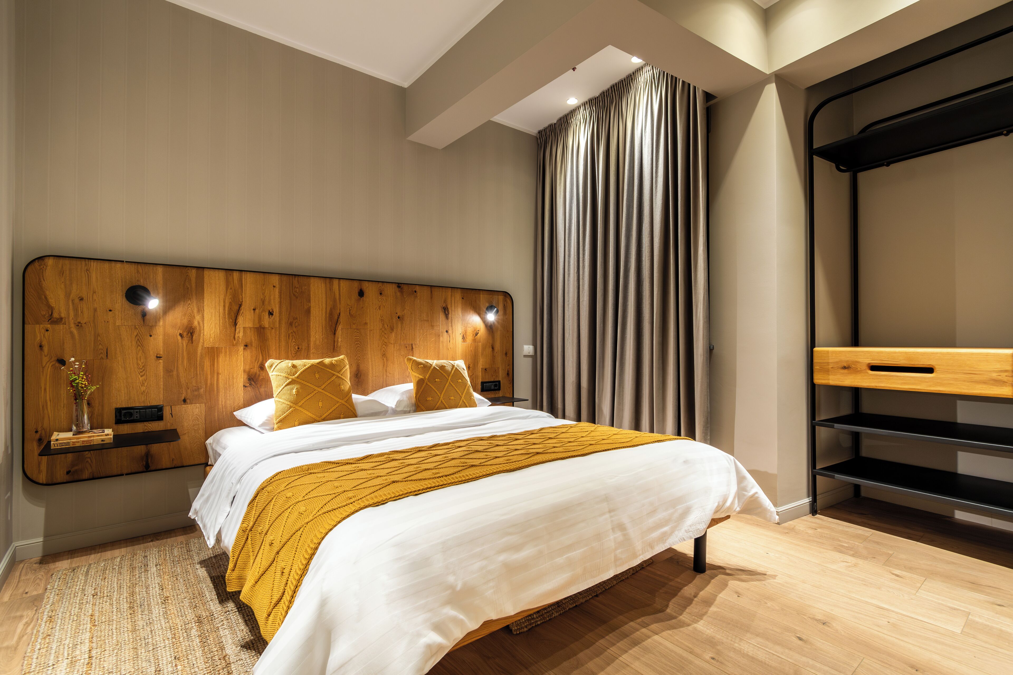 Une chambre moderne avec une tête de lit en bois, un lit recouvert de draps blancs et agrémenté d’un plaid jaune, une table de chevet avec un vase, et une grande fenêtre avec des rideaux.