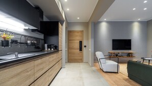 Una cocina moderna con armarios de madera, estufa integrada y encimera negra. Una puerta de madera da acceso a otra habitación. Un sofá verde y una pequeña mesa se encuentran en la sala de estar, con un televisor de pantalla plana montado en la pared.