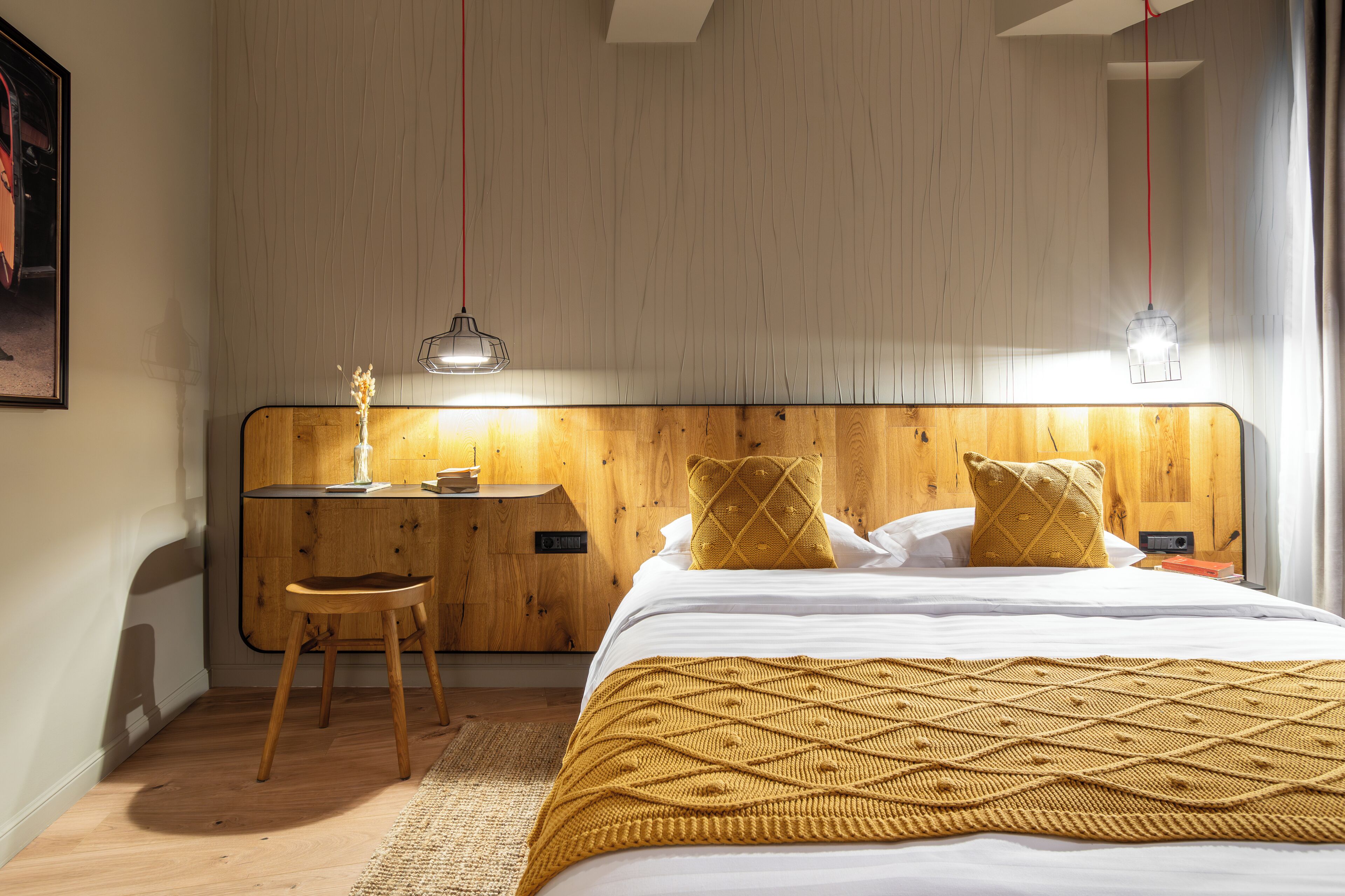 Une chambre à coucher avec une tête de lit en bois, un lit recouvert de draps blancs, un plaid de couleur moutarde, une table de chevet en bois avec une lampe et un petit tabouret.