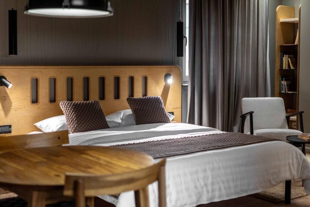Habitación de hotel ordenada, con cabecera de madera, cama con ropa blanca, una mesita redonda y una silla.