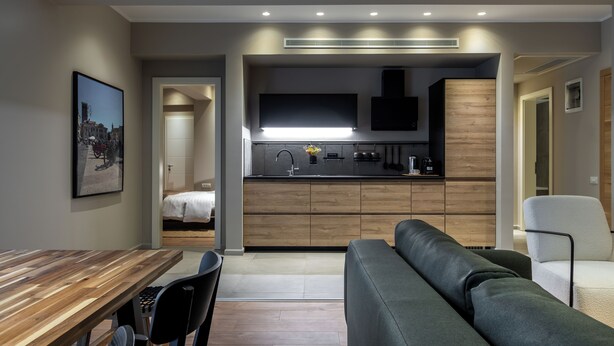 Una cocina moderna con estufa integrada, televisor de pantalla plana y armarios de madera. Cuenta con un comedor con mesa y sillas de madera, y un sofá gris.