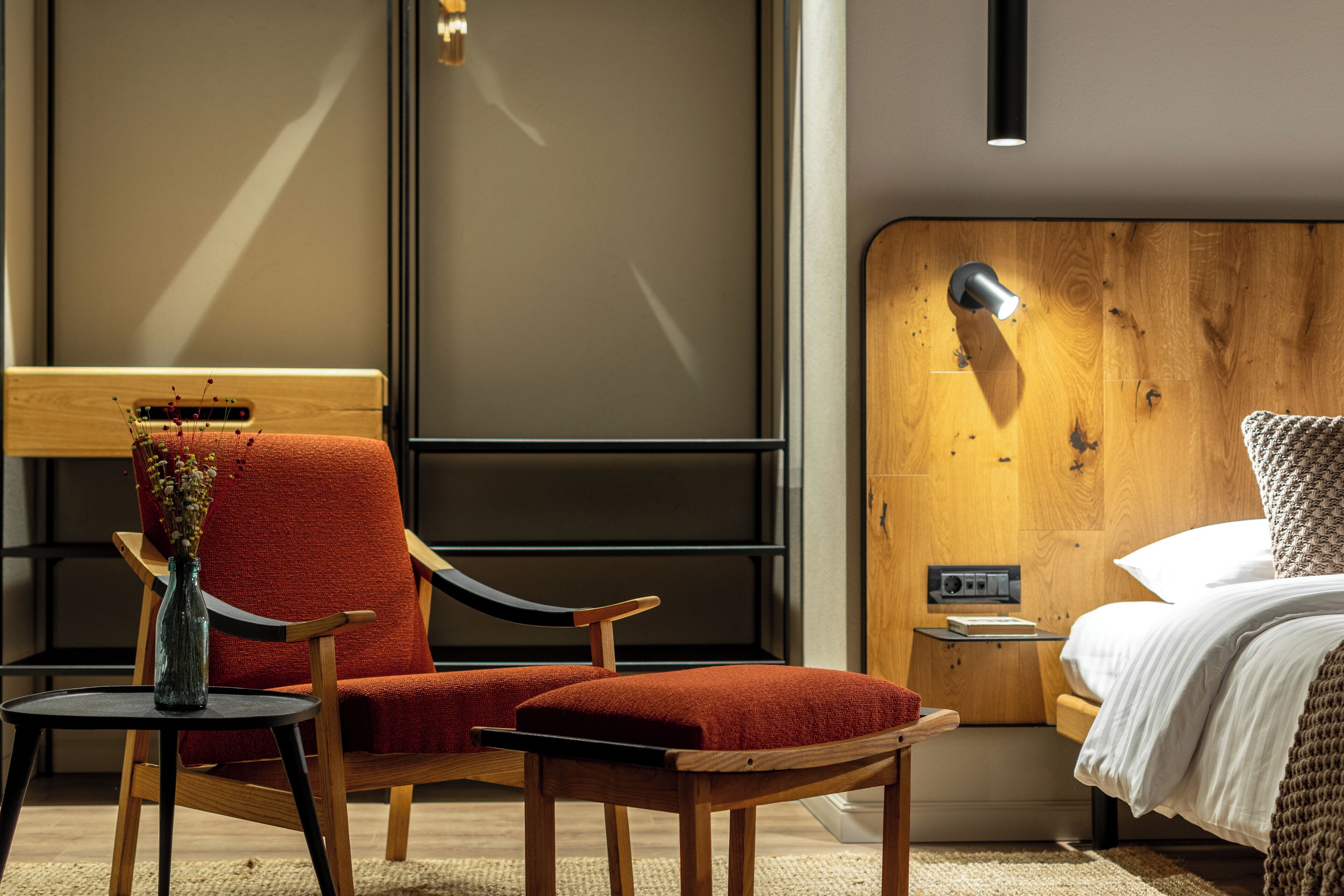 A modern hotel room with a wooden headboard, a red upholstered chair, a small table with a vase, and a bedside lamp.