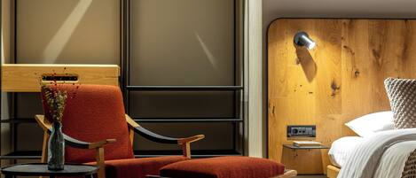 A modern hotel room with a wooden headboard, a red upholstered chair, a small table with a vase, and a bedside lamp.