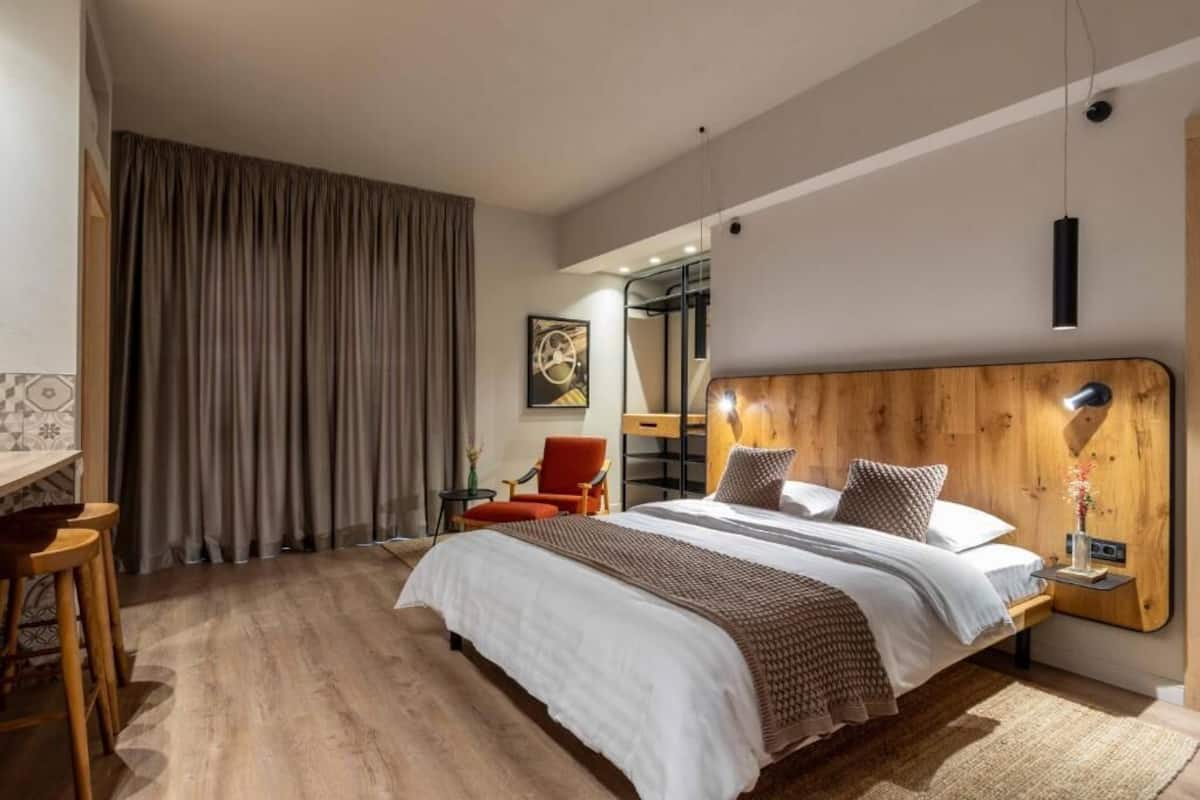 Une chambre d’hôtel moderne avec un grand lit, une tête de lit en bois, un bureau avec une chaise et une grande fenêtre avec des rideaux.