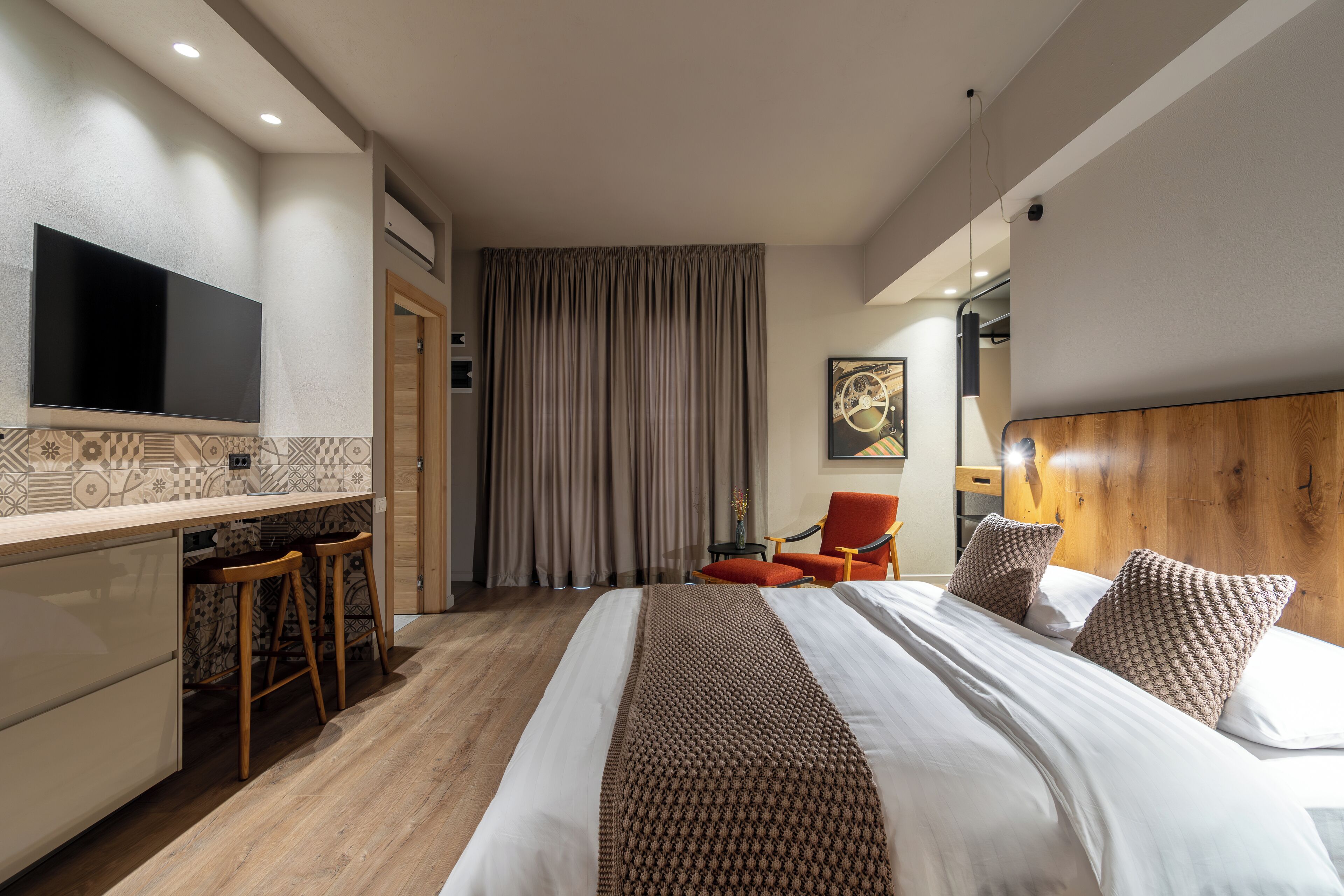 A modern hotel room with a large bed, a desk with two stools, a flat-screen TV mounted on the wall, and a wooden headboard.