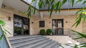 Una entrada de edificio moderno con una escalera que conduce a un balcón, rodeada de vegetación y con fachada de ladrillo.