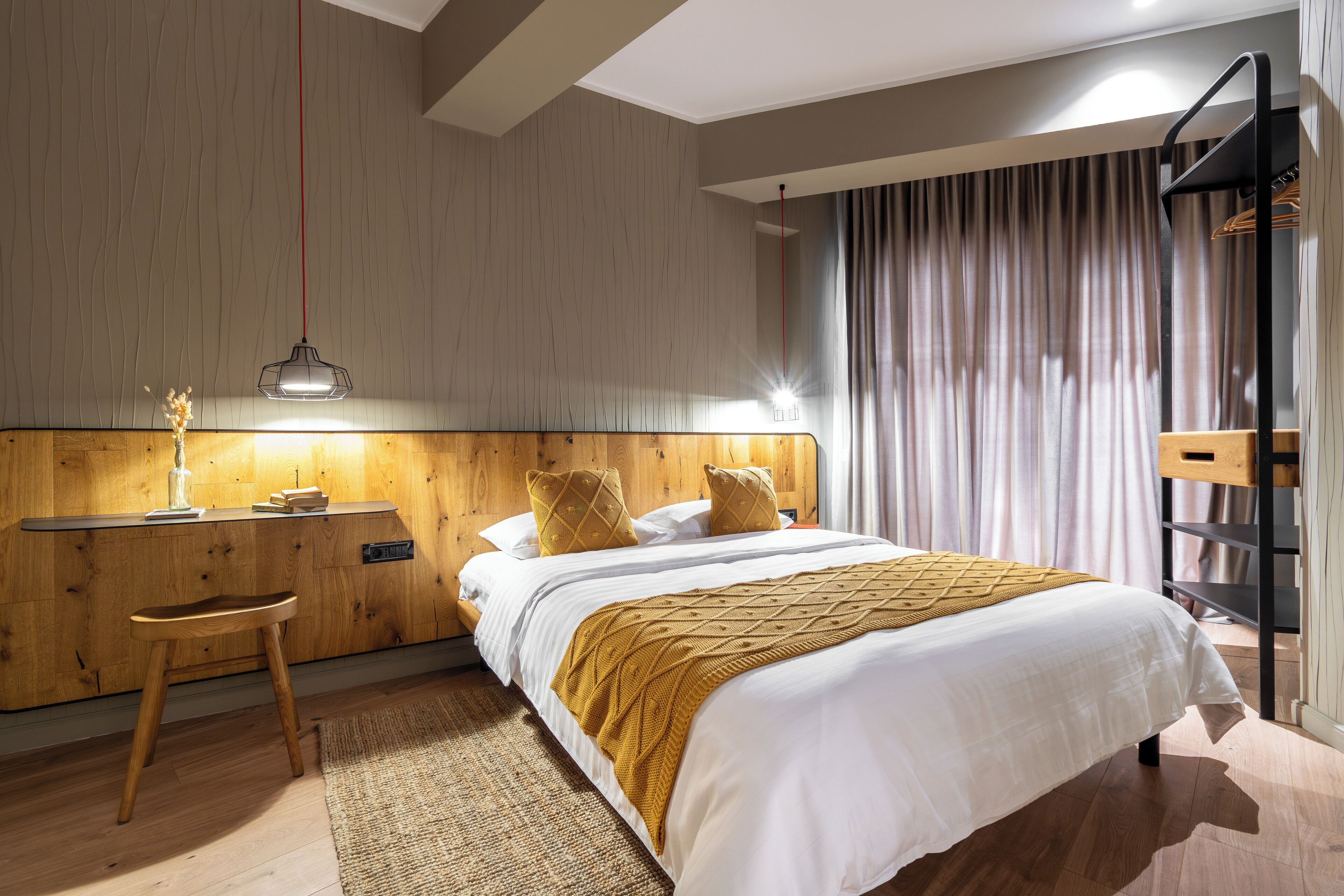 A modern bedroom with a wooden headboard, a bed with white linens and a mustard-colored throw, a wooden nightstand, a desk with a lamp, and a built-in wardrobe.