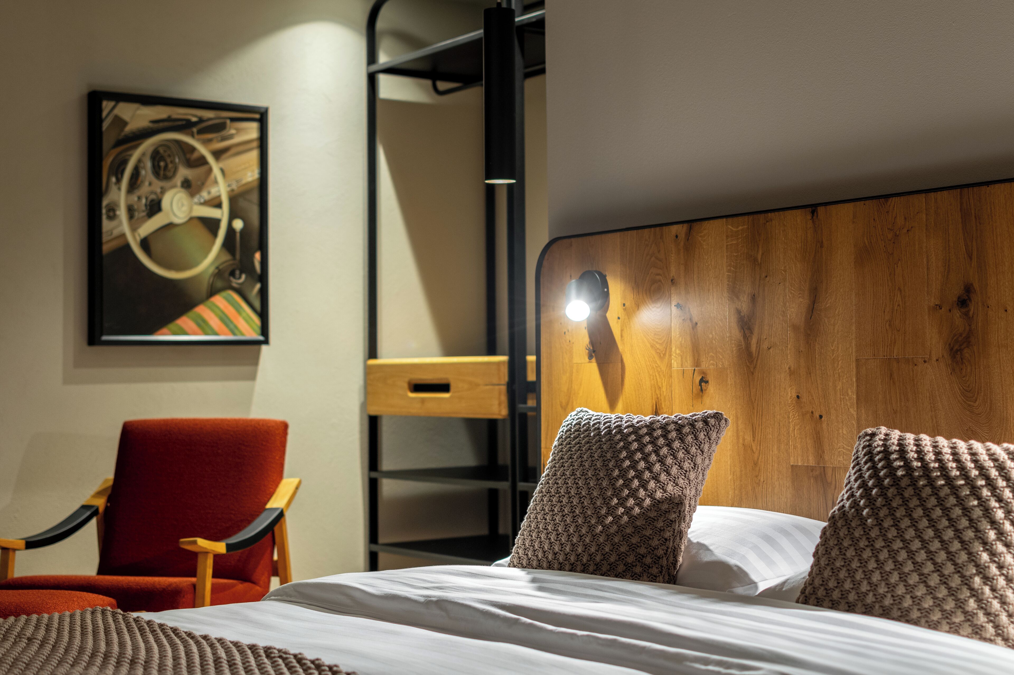 A bedroom with a bed, pillows, a chair, a wooden headboard, a shelf, and a wall-mounted artwork.