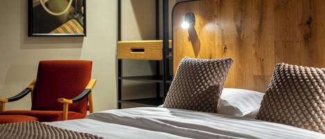 A bedroom with a bed, pillows, a chair, a wooden headboard, a shelf, and a wall-mounted artwork.