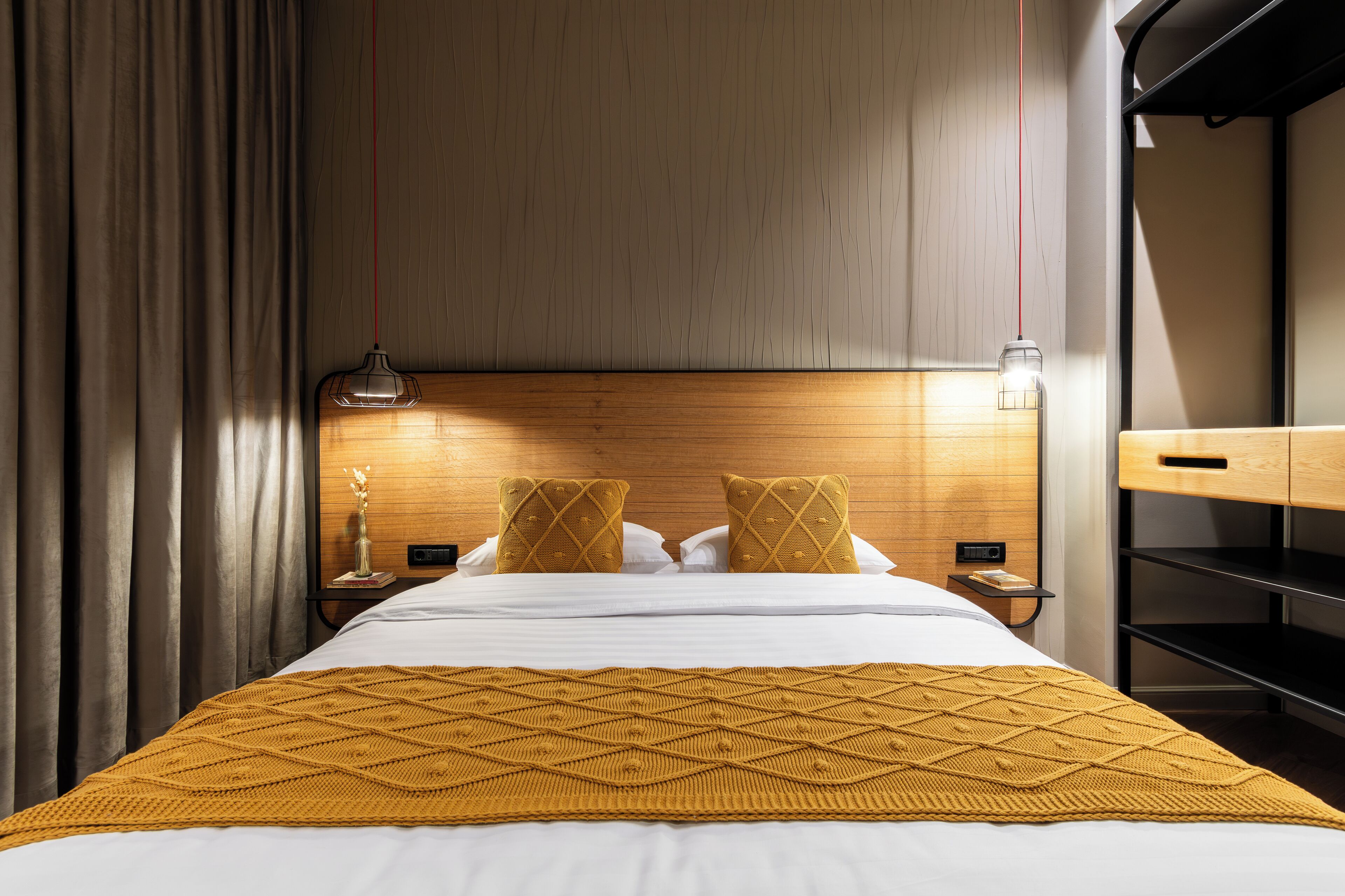 A hotel room with a wooden headboard, a bed with white linens, a mustard-colored throw blanket, and a nightstand with a lamp and a small table.