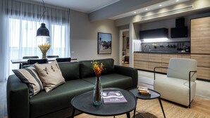 Un salón moderno con un sofá verde, una mesa de centro redonda con un jarrón y una revista, un sillón blanco y una zona de cocina al fondo.