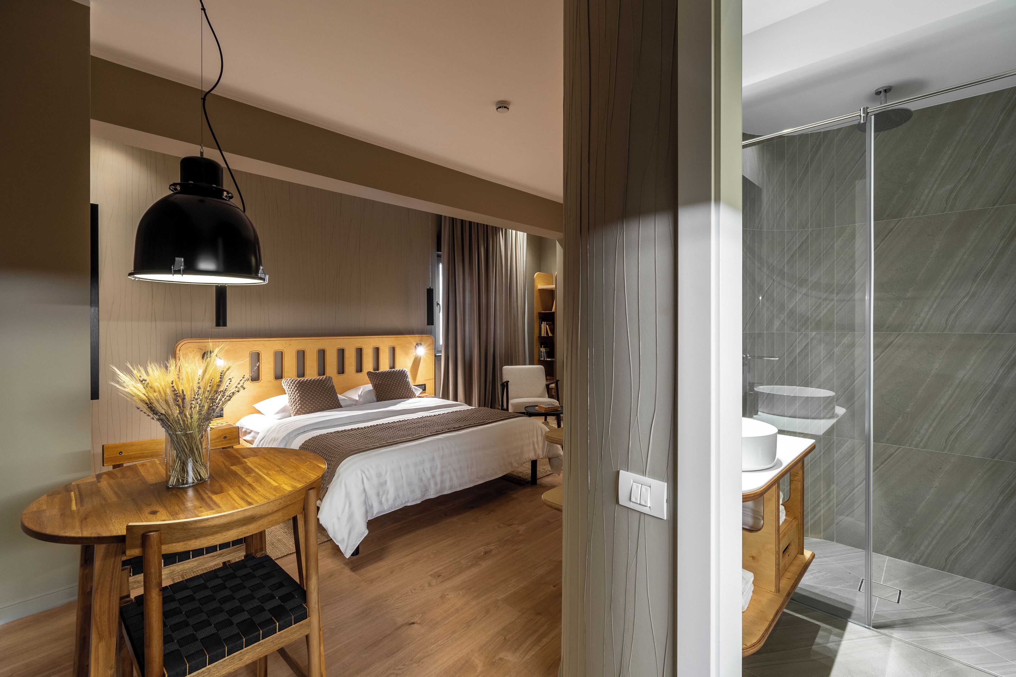 A modern hotel room with a wooden bed, a bedside table with a vase, a wooden chair, a desk, and a shower area with a glass door.