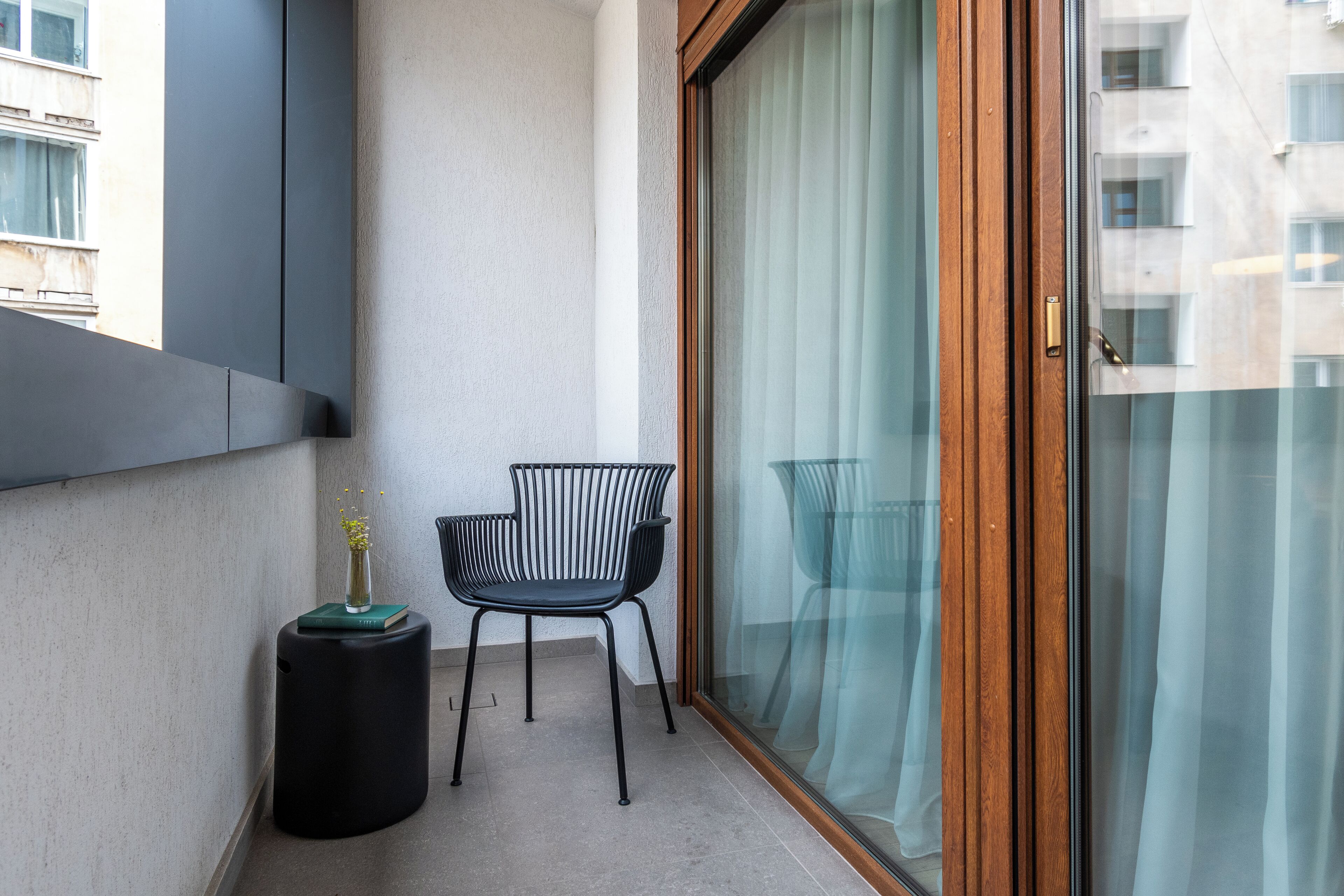 A balcony with a black chair, a small round table with a vase, and a sliding glass door with curtains.