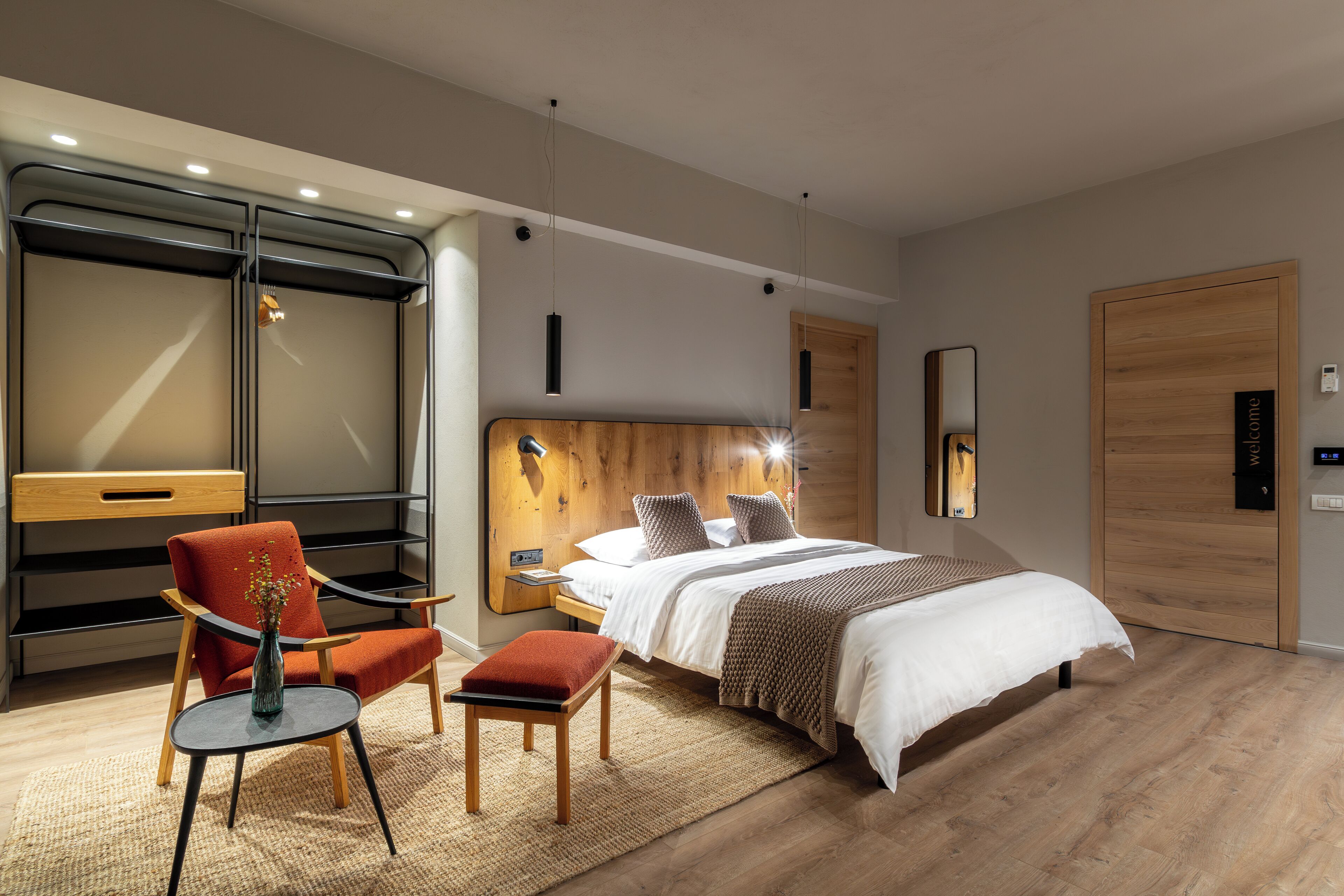 A modern hotel room with a large bed, a wooden headboard, a red armchair, a small table with a vase, and a large wardrobe.