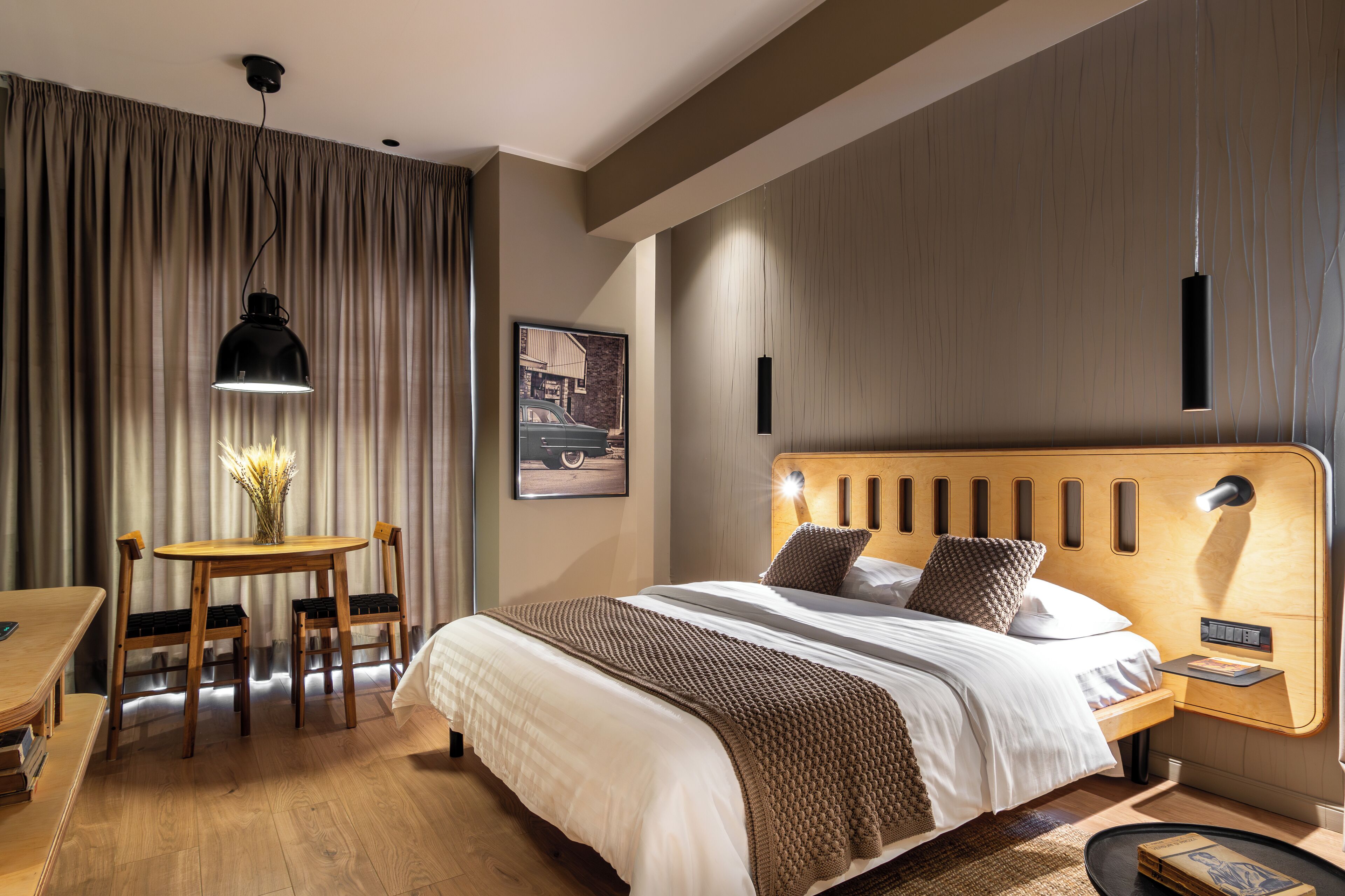 Una habitación de hotel moderna con un cabecero de madera, una cama grande, una mesita de noche, un escritorio con sillas y un ventanal con cortinas.