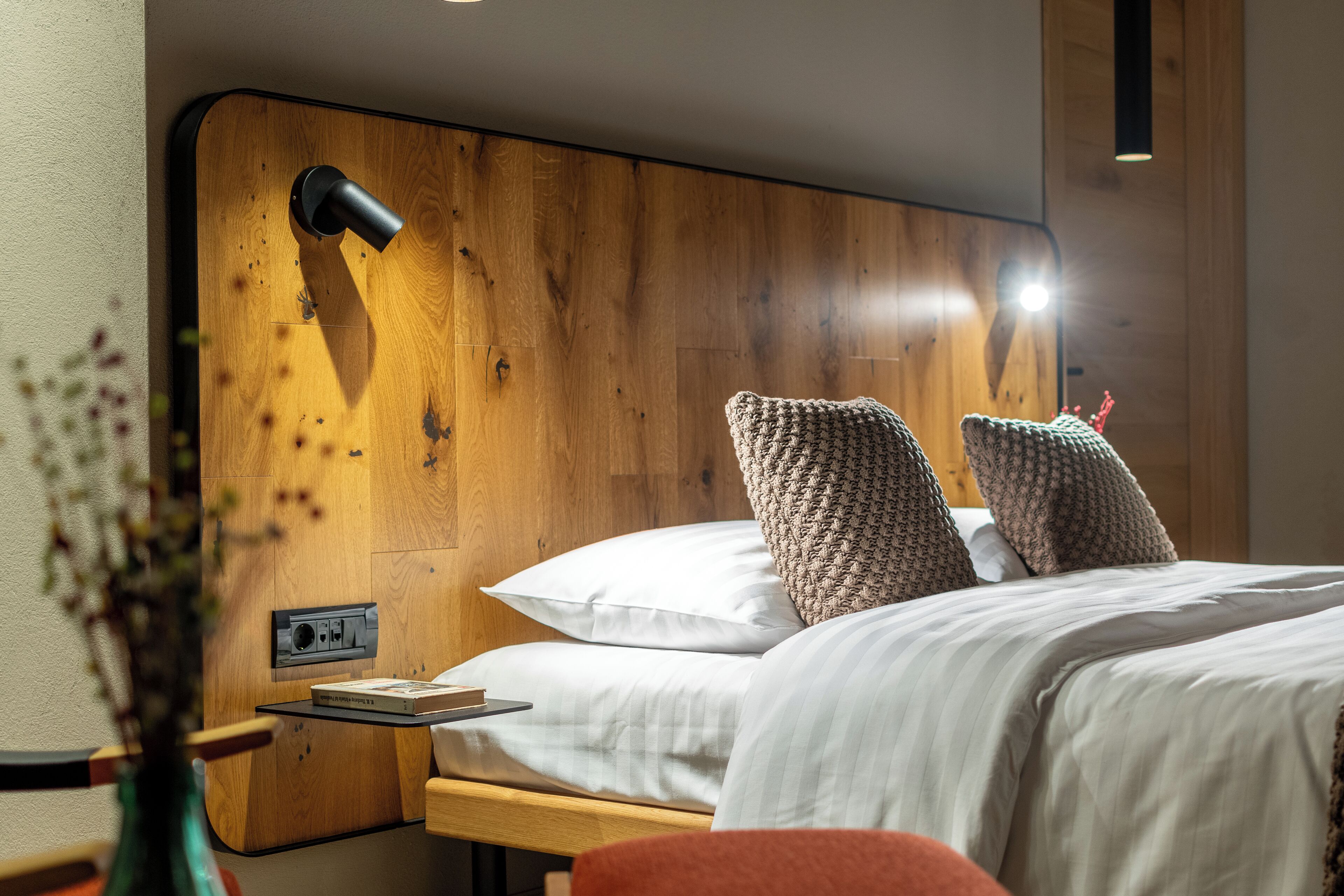 A hotel room with a wooden headboard, a bedside table with a lamp, and two pillows.