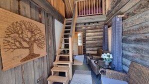 A log cabin interior with a wooden staircase, a woven rug with a tree design, wicker furniture, and a small table with a vase of flowers. - Kaunisvirta villa for 10 with beautiful views. Pet-Friendly! (Pyhtään kirkonkylä)
