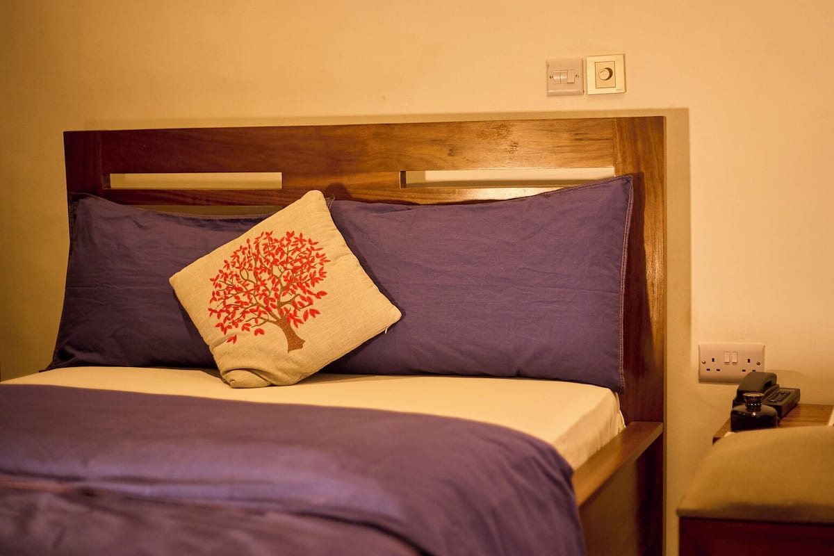 Ein ordentlich bezogenes Bett mit einem hölzernen Kopfteil, einem violetten Kopfkissen und einem beigefarbenen Kopfkissen mit einem roten Baumdesign.