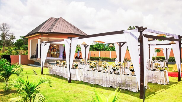An outdoor wedding venue with a covered seating area, a stage, and a building in the background. - JLK Guest House & Events Centre (Koforidua)