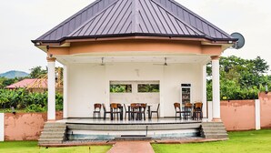 A covered outdoor seating area with tables and chairs, a pathway leading to it, and a building with a dark roof in the background. - JLK Guest House & Events Centre (Koforidua)