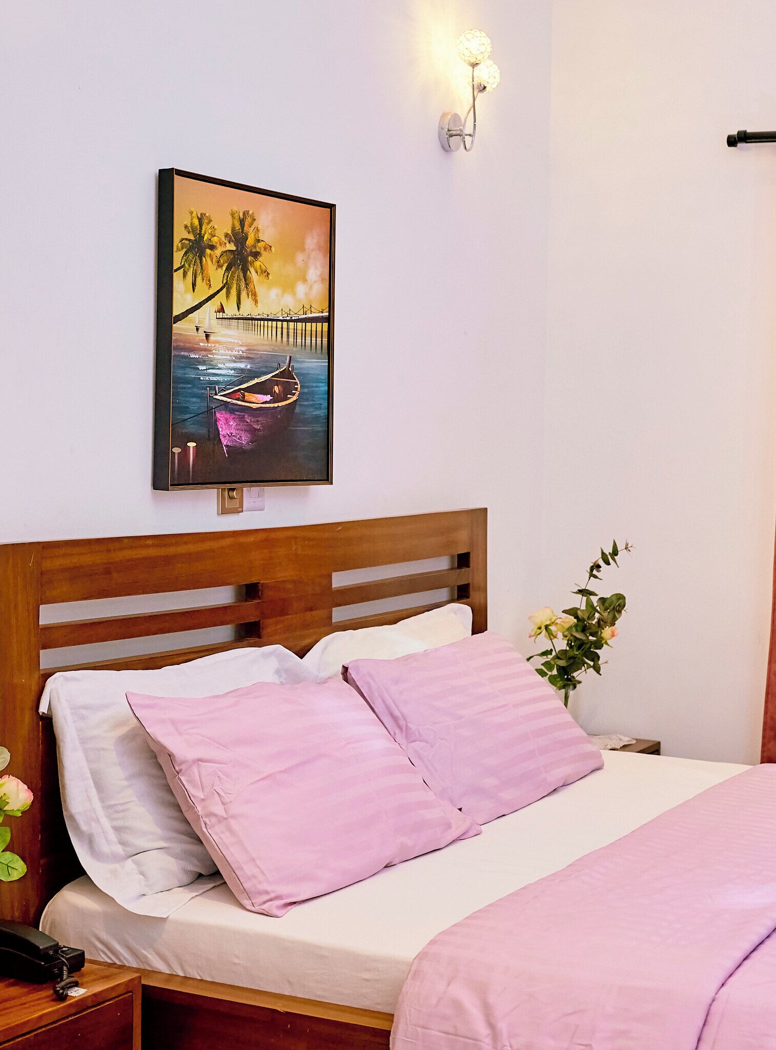 A hotel room with a wooden headboard, a bed with pink and white bedding, a framed picture of a beach scene, a bedside table with a phone, and a wall-mounted light fixture.